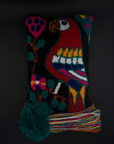 A rectangular hand-hooked clutch with a black background and a colorful parrot and floral design, featuring a large dark green pom-pom and multicolored tassel on the left side, set against a dark background.