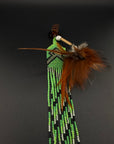 A South American style beaded brooch or pin featuring a turquoise stone, brown feathers, and long strands of green, black, and white beads, displayed on a dark gray background.