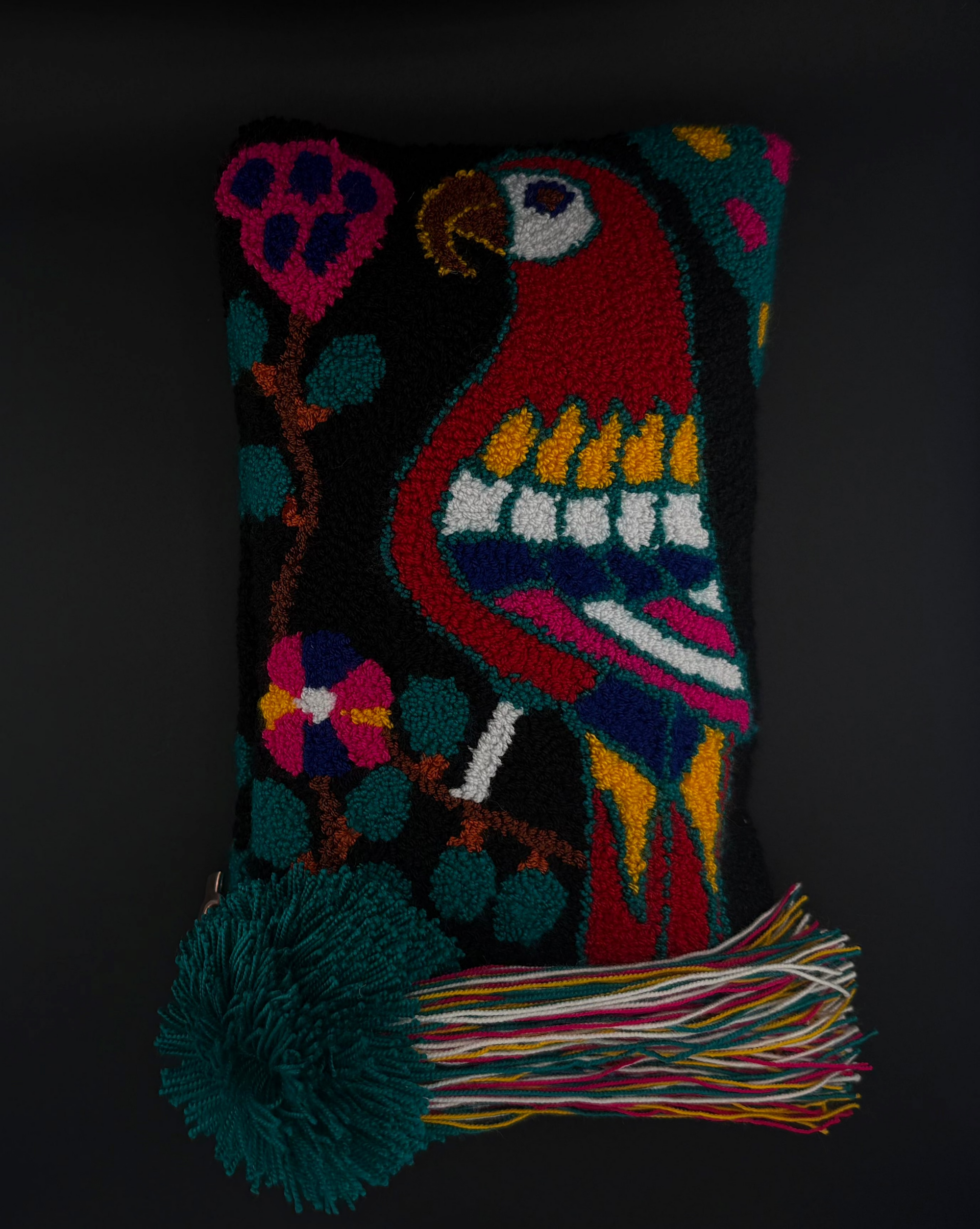 A rectangular hand-hooked clutch with a black background and a colorful parrot and floral design, featuring a large dark green pom-pom and multicolored tassel on the left side, set against a dark background.
