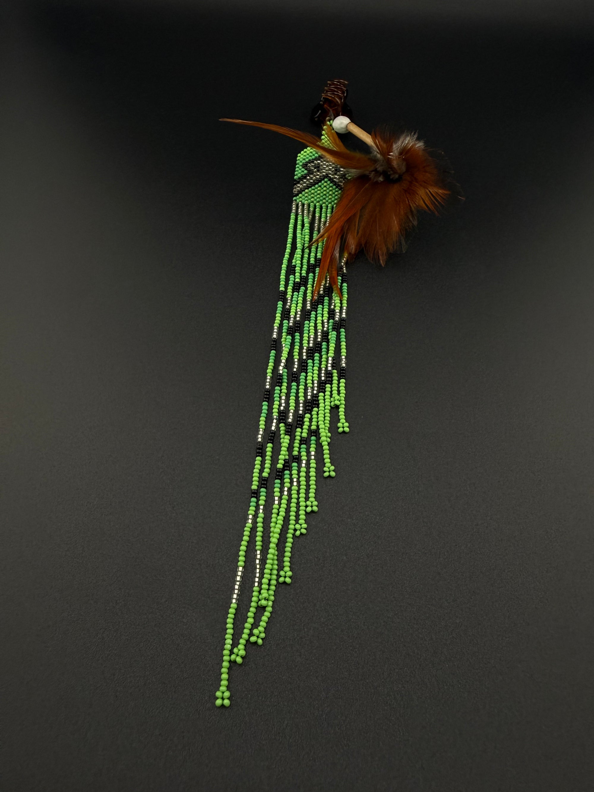 A South American style beaded brooch or pin featuring a turquoise stone, brown feathers, and long strands of green, black, and white beads, displayed on a dark gray background.