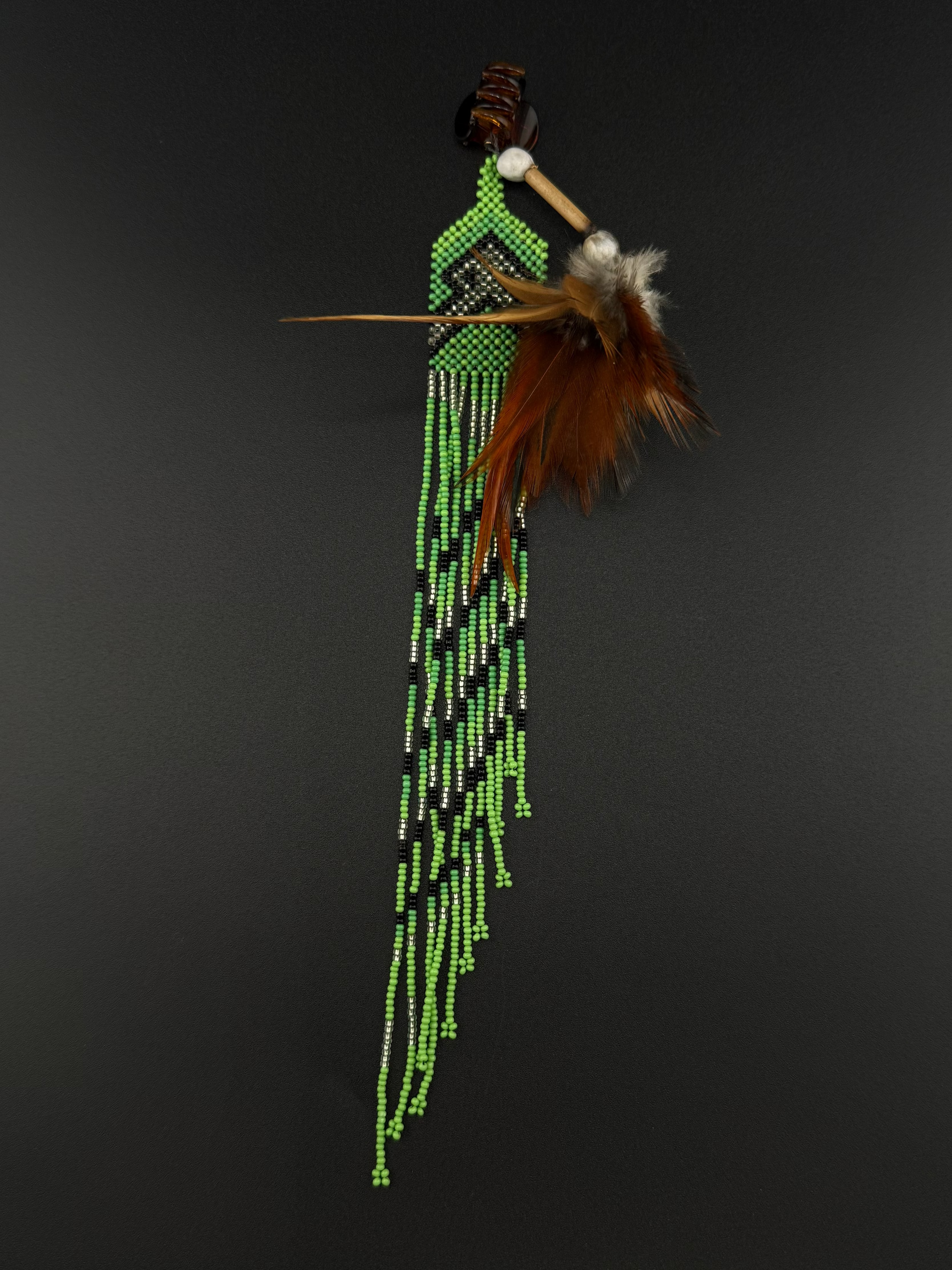 A South American style beaded feather ornament featuring bright green, black, and white beads, a brown wooden shaft, and brown feathers, displayed against a dark gray background.