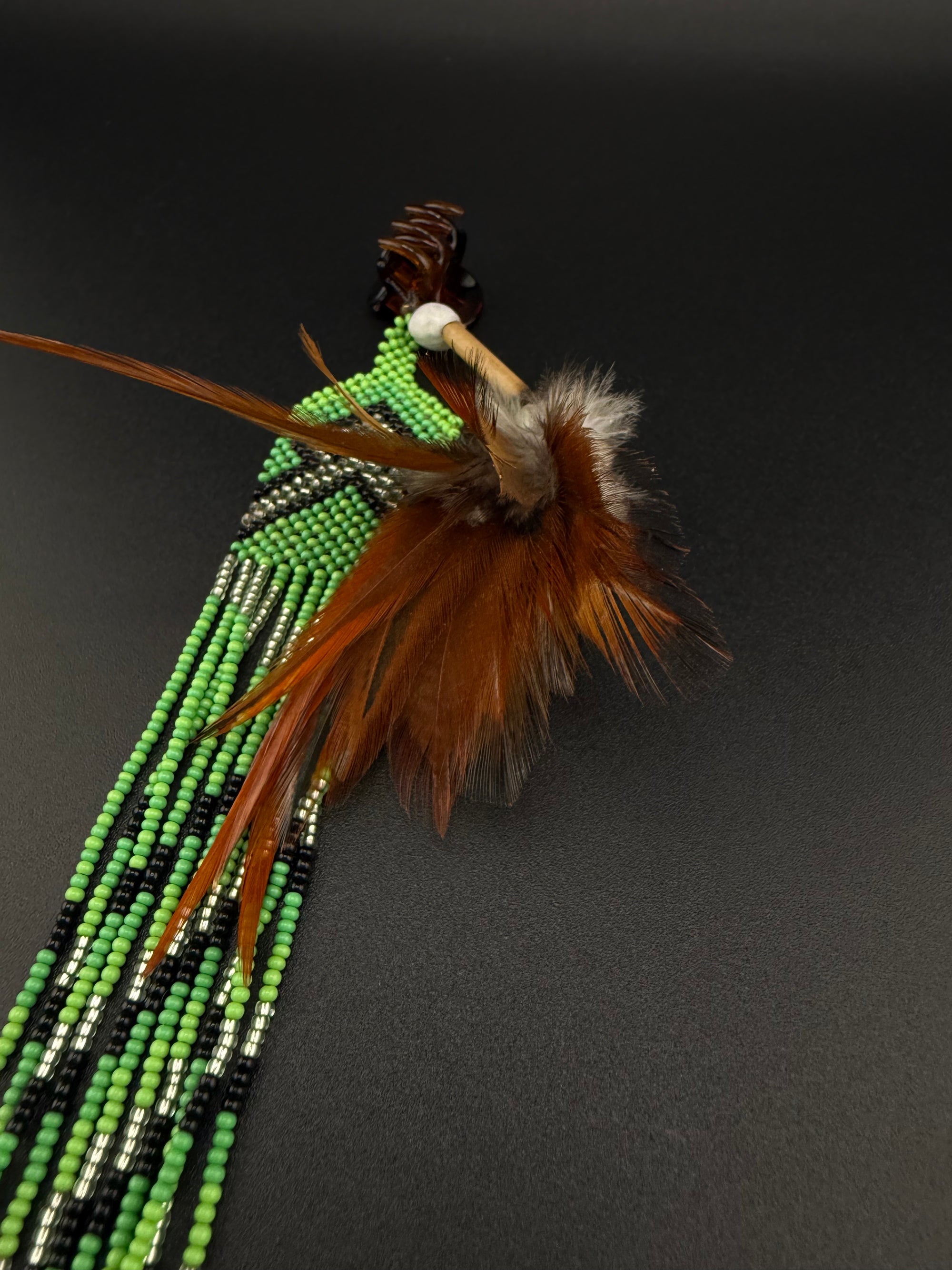 A South American style beaded brooch or pin featuring a turquoise stone, brown feathers, and long strands of green, black, and white beads, displayed on a dark gray background.
