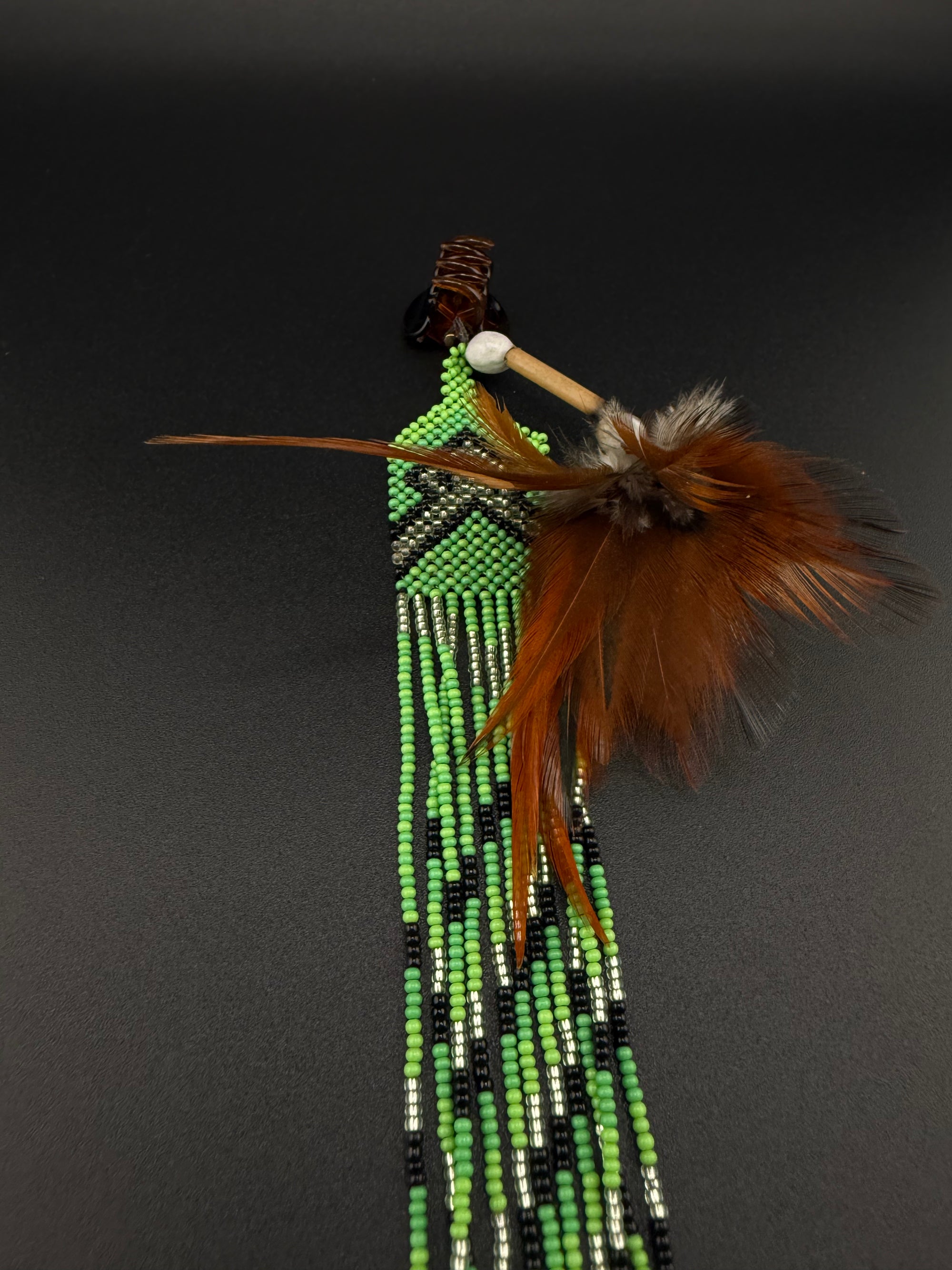 A South American style beaded brooch or pin featuring a turquoise stone, brown feathers, and long strands of green, black, and white beads, displayed on a dark gray background.