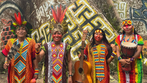 Four people standing in front of a massive tree trunk in a forest, wearing colorful traditional-style garments with geometric patterns and feathered headpieces, with one holding a guitar and another holding a drum.